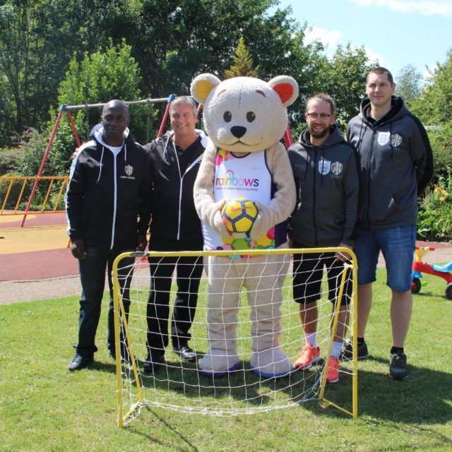 Four men standing with Rainbows mascot, Bow Bear, who is holding a football
