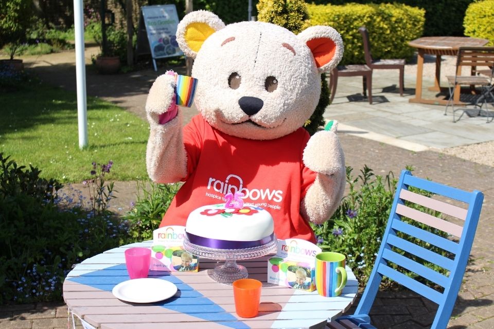 Rainbows mascot Bow Bear holding a cup with thumbs up, behind a cake