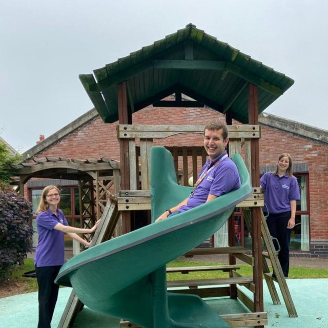Three smiling people in purple tshirts around a green slide with a wooden frame.